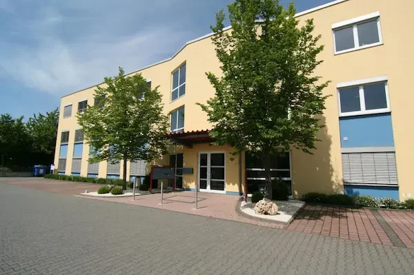 Gelbes Geschäftsgebäude mit Bäumen und Parkplatz. Eingangstüren sind sichtbar.